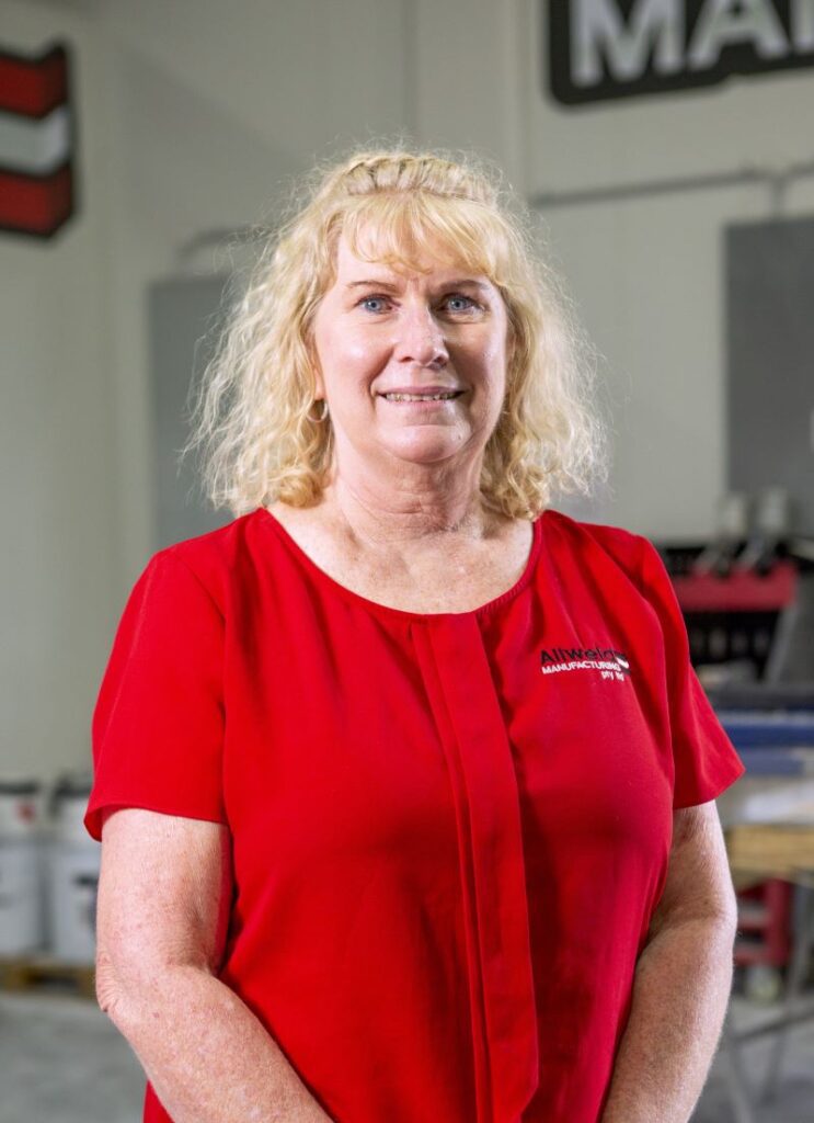 A woman with shoulder-length blonde hair, wearing a bright red blouse, stands indoors in a workspace with equipment and supplies visible in the background.