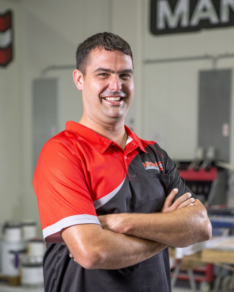 A man with short dark hair in a red and black polo shirt stands with arms crossed and smiles at the camera. The background shows workshop equipment and paint cans.