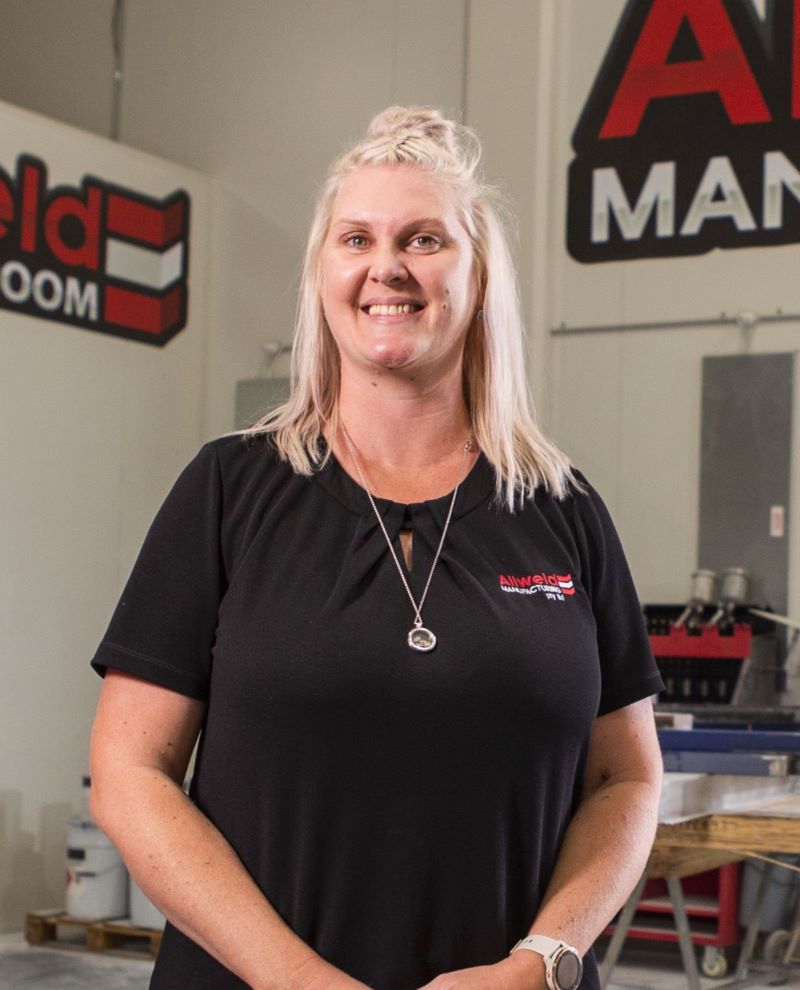 A person with long blonde hair, wearing a black shirt with a logo, stands in an industrial-looking room with signs in the background. They are smiling and have a pendant necklace and a watch.
