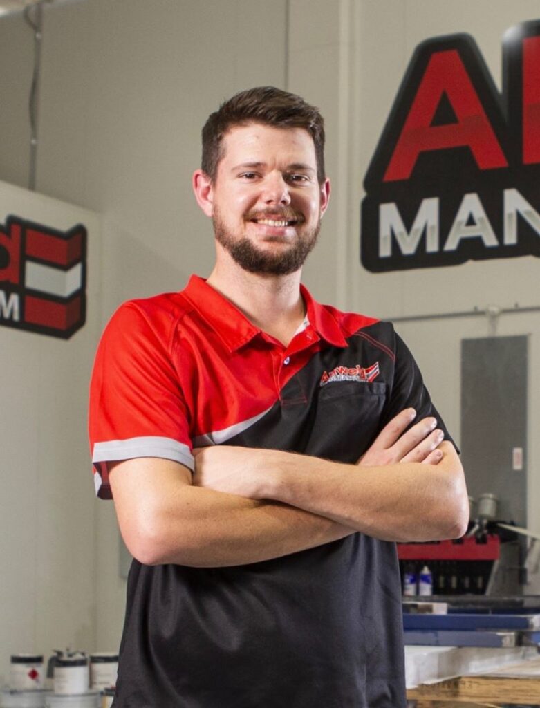 A man with a beard stands confidently with arms crossed, wearing a red and black polo shirt in a workshop. There are industrial tools and equipment in the background, along with a large sign on the wall.