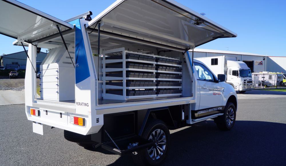 A white utility truck with storage compartments and shelves in the open cargo area, showcasing vehicle fitouts and modifications, parked on asphalt near industrial buildings with both side and rear canopy doors open.