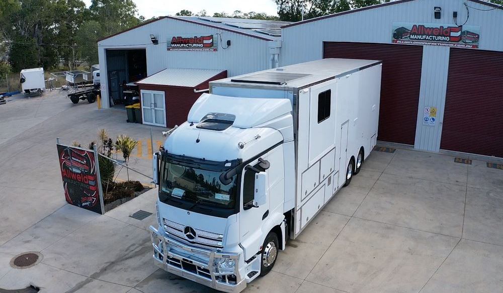 A large white Mercedes-Benz truck, designed for custom transporters and trailers, is parked outside a building with Allweld Manufacturing signage. The clean industrial area features a few other vehicles and trees in the background.