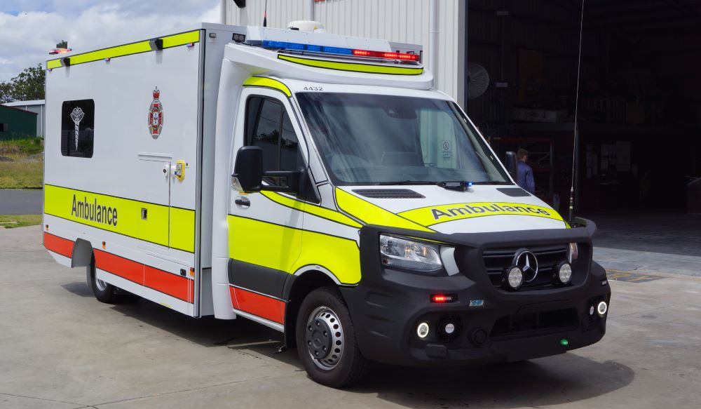 A modern ambulance, part of the emergency service vehicles fleet, with yellow and green reflective markings, is parked outside a building. The vehicle displays flashing lights and has “Ambulance” written on the hood and side.