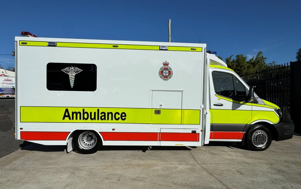 A white and yellow ambulance with red accents is parked on a paved surface under a clear blue sky. The vehicle has the word Ambulance, a medical symbol, and an official crest on its side.