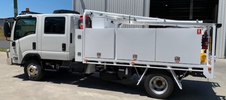 A white utility truck with multiple storage compartments is parked outside a large industrial building on a sunny day.