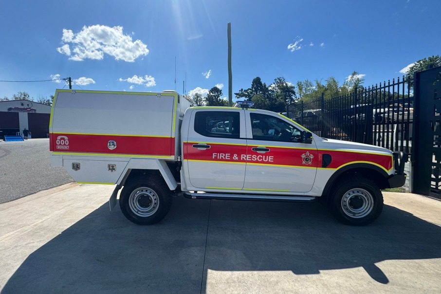 A white Fire & Rescue utility truck with red and yellow stripes is parked on a concrete surface beside a black metal fence under a clear blue sky.