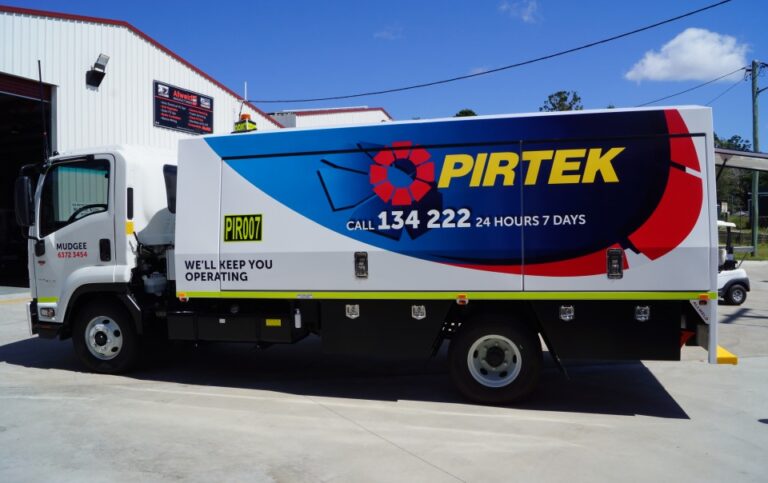 A white Pirtek service truck with bold blue, red, and yellow branding and contact number is parked outside an industrial building on a sunny day. The side of the truck reads WELL KEEP YOU OPERATING.