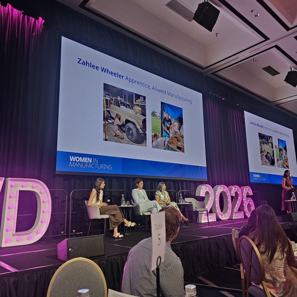 A panel of women sits on stage at a conference. A large screen displays Zahlee Wheeler, Apprentice, Allied Manufacturing with photos of her at work. Illuminated “WIM” and “2026” signs are visible on stage.