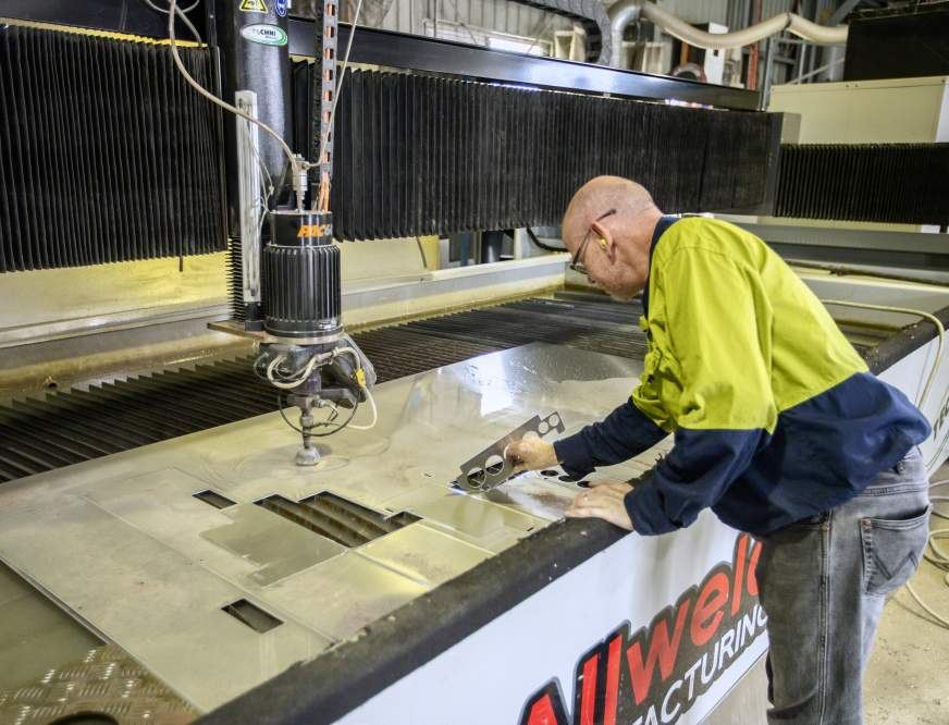 A person in a high-visibility jacket operates a large industrial cutting machine, inspecting a metal cutout piece at Allweld Manufacturing’s facility.
