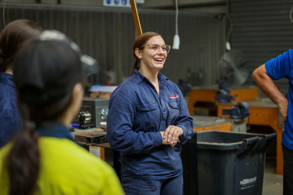 A woman in safety glasses and a blue work jumpsuit stands smiling and talking in a workshop, with two other people visible in the foreground. Workbenches and tools are in the background.