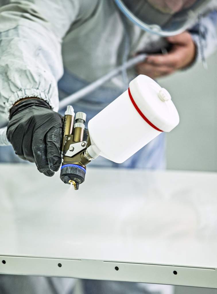 A person wearing protective gear and black gloves holds a spray paint gun, applying a white coating to a flat surface.