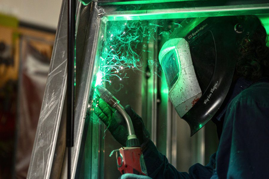 A person wearing a welding helmet and protective clothing uses a welding torch, with green light and smoke visible around the work area.