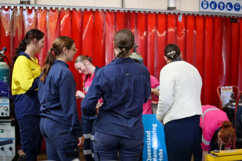 A group of women in blue and pink work uniforms stand and chat in front of a red curtain in an industrial or workshop setting. Some face away, while others are engaged in conversation or activities.