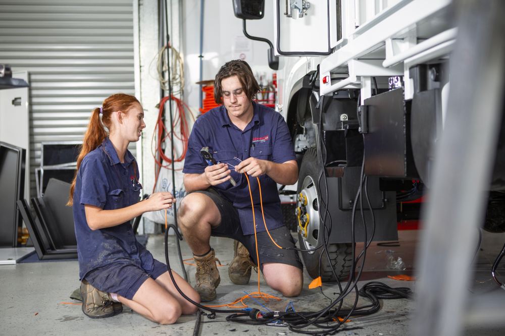 Two mechanics in blue uniforms kneel beside a white vehicle in a garage, working together with electrical wires and tools. Coiled cables and equipment are visible around them.