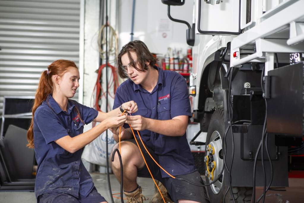Two young mechanics in blue uniforms work together, handling electrical wires beside a white truck in a garage filled with tools—showcasing the hands-on expertise found at Allweld Manufacturing.