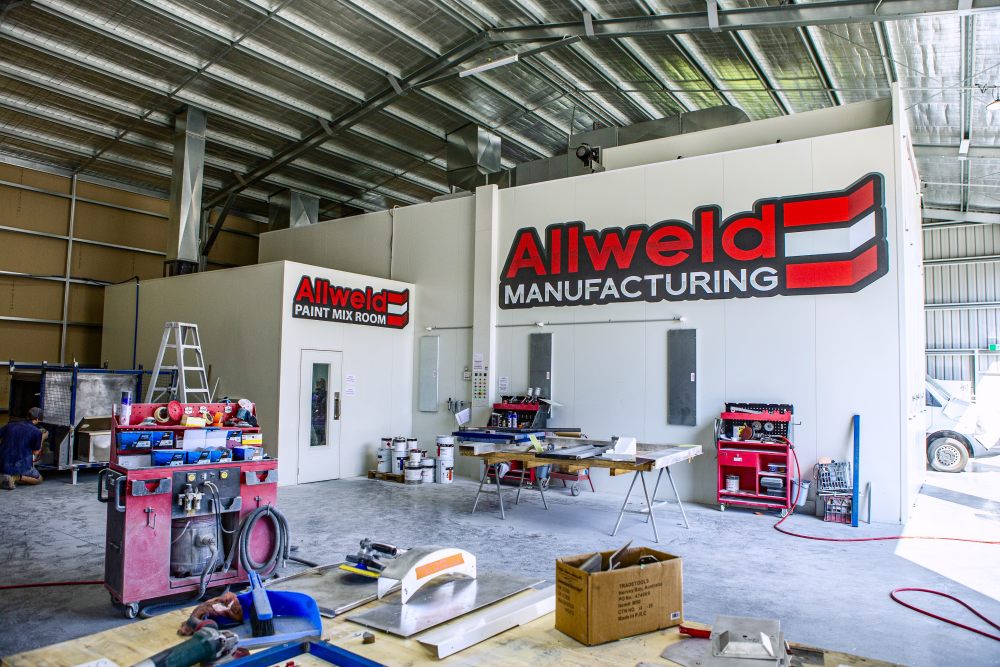 Interior of a large industrial workshop with tools, equipment, and workbenches. A white structure displays signs reading "About Allweld Manufacturing" and "Paint Mix Room." The area appears organized but actively used.