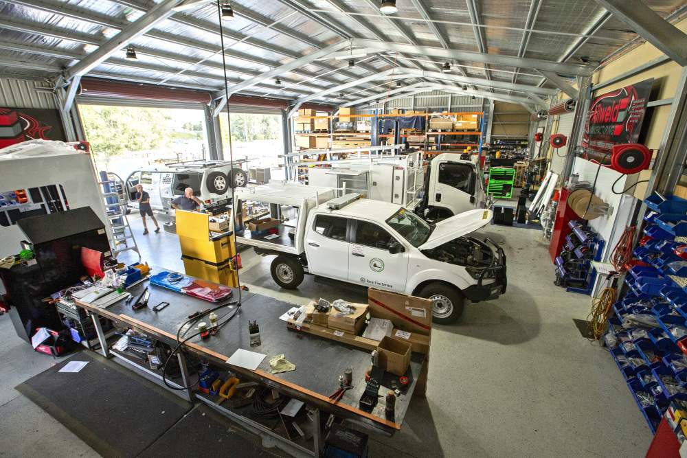 A spacious workshop with high ceilings, several vehicles being serviced, workbenches covered with tools and parts, shelves with equipment, and people working on different automotive tasks.